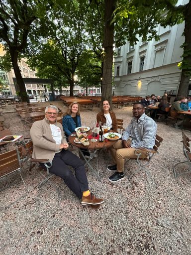 Vier Personen sitzen an einem Tisch im Biergarten in München, umgeben von Bäumen und einer belebten Atmosphäre.