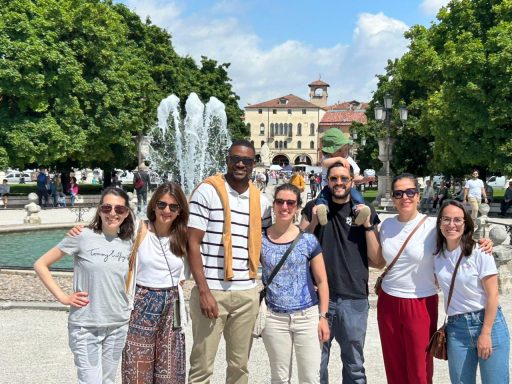 Gruppe von acht Personen vor einem Brunnen in einem Park mit Bäumen und Gebäuden im Hintergrund.